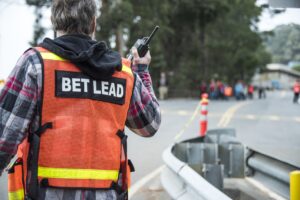 A person with their back to us wears an orange vest that says "BET Lead". They are holding up a radio as people evacuate in the distance. 